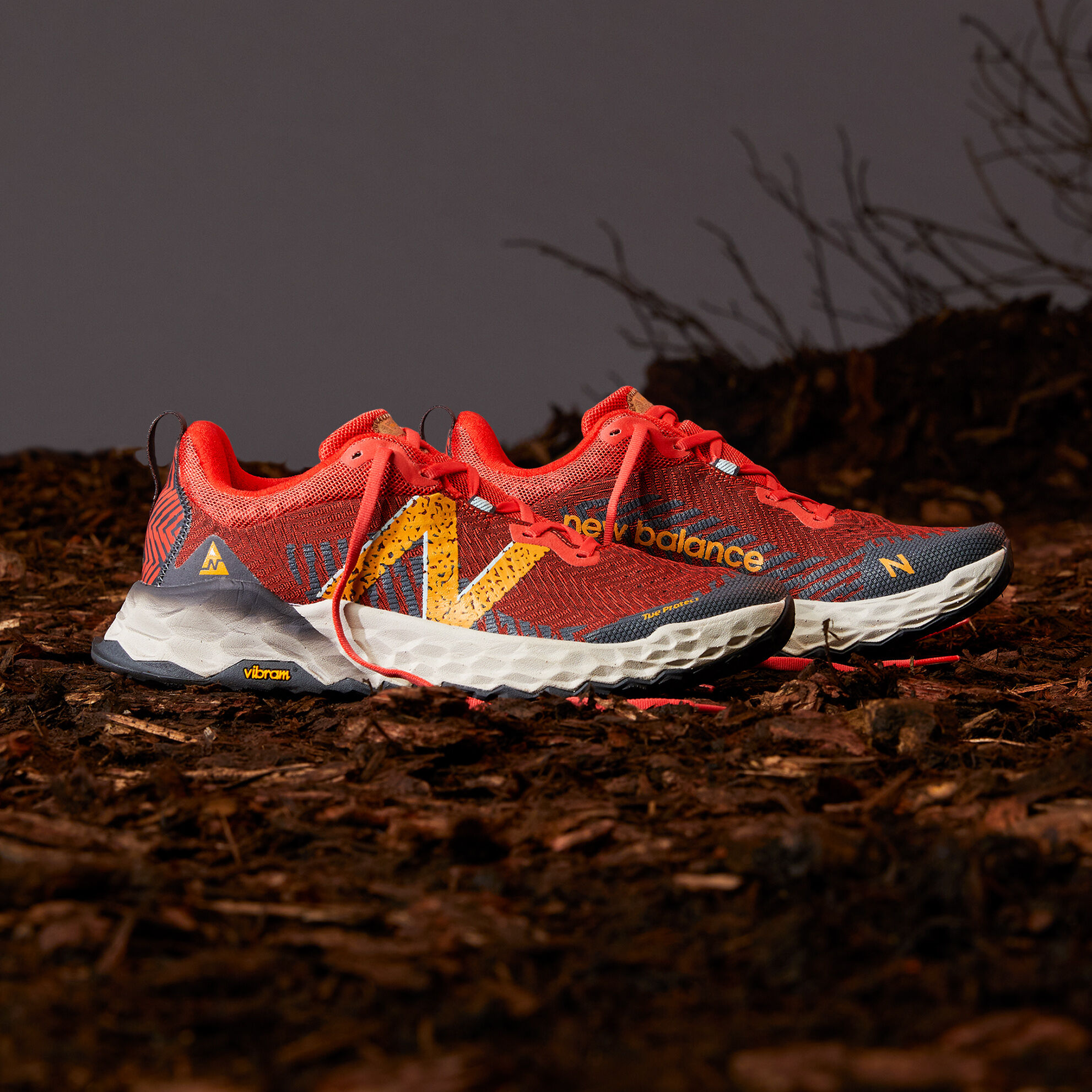 A pair of bright orange men’s trail shoes on dirt and sticks with a dark grey background. 