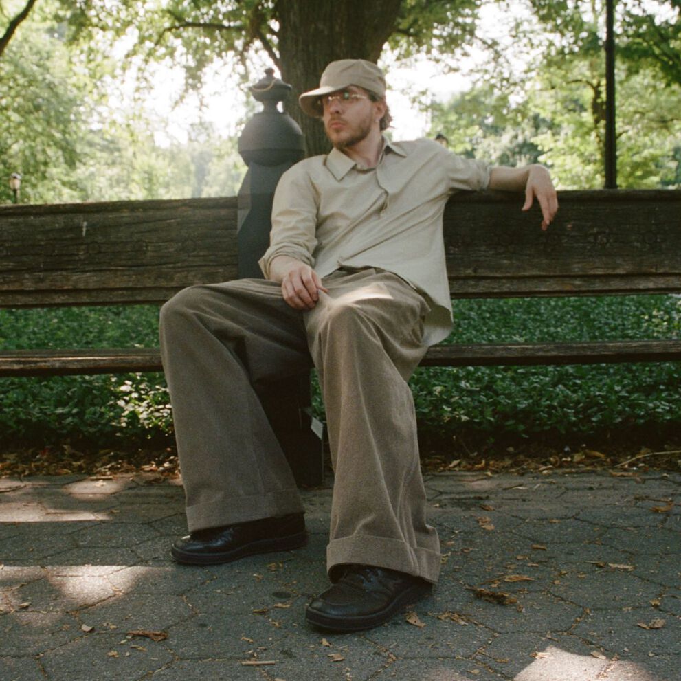 Jack Harlow reclinado en un banco en un parque, usando zapatillas New Balance color marr&oacute;n oscuro.